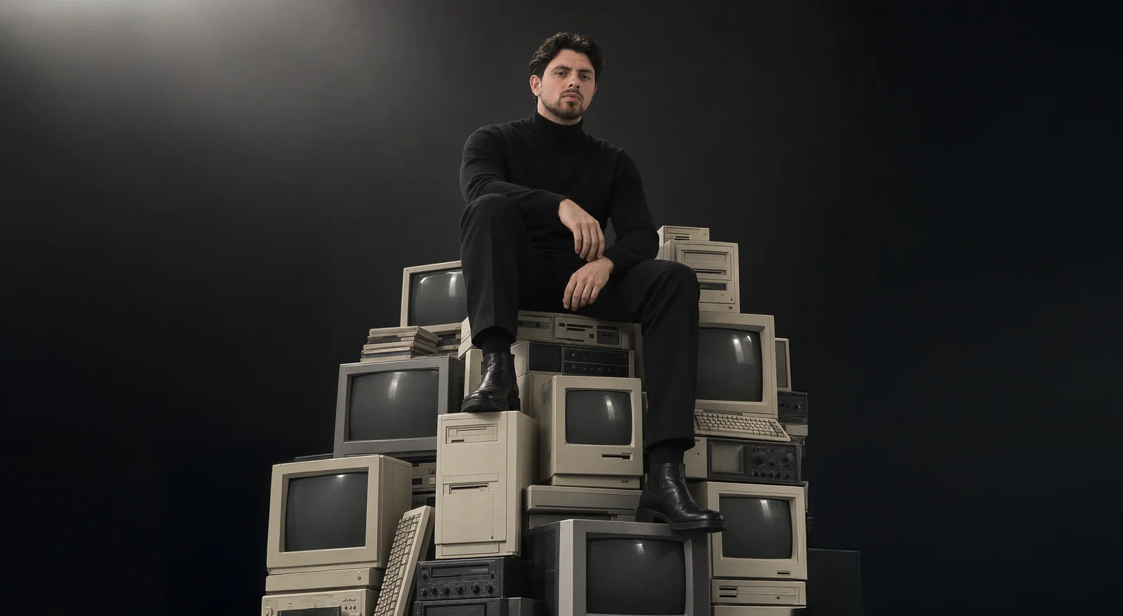 Alain Tural seated on a stack of vintage computers, landscape frame